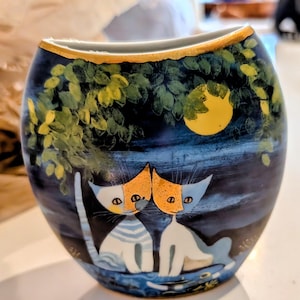 May include: A ceramic vase with a dark blue background featuring a painted scene of two stylized cats under a tree and a yellow moon. The cats have orange and white markings. The rim of the vase has a gold trim.