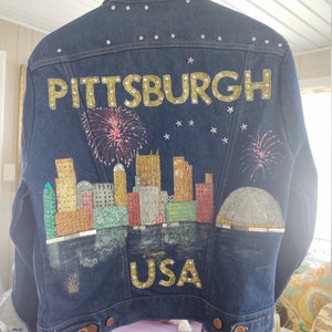 May include: A blue denim jacket with a gold glitter design of the Pittsburgh skyline and the words "PITTSBURGH USA".
