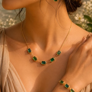 May include: A gold-toned jewelry set consisting of a necklace, a bracelet, and a single earring, each featuring rectangular, emerald-green faceted stones set in gold-colored metal frames. The necklace and bracelet are composed of linked stone settings.