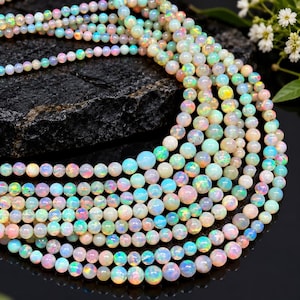 May include: A collection of smooth, round opal beads arranged in multiple strands. The beads exhibit a translucent white base with iridescent flashes of blue, green, pink, and yellow light. The strands are layered, showcasing a variety of bead sizes.