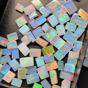 Könnte beinhalten: Eine Sammlung von schillernden Opal-Edelsteinen in verschiedenen Größen und Formen, die in einem schwarzen Tablett präsentiert werden. Die Opale haben eine milchig weiße Basis mit grün, blau und orangefarbenen Blitzen.