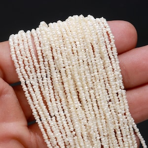 Może przedstawiać: Sznur białych pereł słodkowodnych, każda perła jest mała i o nieregularnym kształcie. Perły są nawleczone na cienki sznurek i mają średnicę około 2 mm.