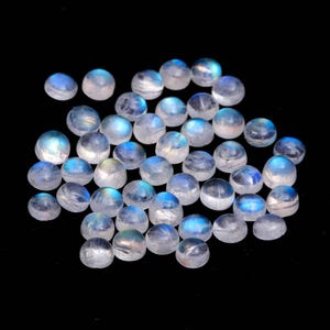 Könnte beinhalten: Eine Sammlung polierter, runder Mondstein-Cabochons. Die Edelsteine sind durchscheinend mit einer milchig-weißen Basis und weisen eine blaue Adulareszenz auf. Die Steine sind auf einem schwarzen Hintergrund verstreut, was ihre schillernde Qualität zeigt.