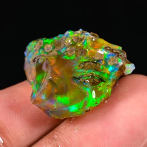 Op de afbeelding: Een levendige, ruwe opaal edelsteen met een mix van kleuren. De steen toont flitsen van groene, blauwe en oranje tinten. De ruwe textuur en natuurlijke vorm van de opaal zijn zichtbaar. De edelsteen wordt in een hand gehouden.
