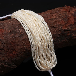 Op de afbeelding: Een streng witte zoetwaterparels, geregen aan een wit koord. De parels zijn klein en uniform van grootte, en ze zijn gerangschikt in een nette, gelijkmatige rij. De parels worden getoond op een stuk bruin hout.