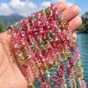 Op de afbeelding: Meerdere strengen gefacetteerde, druppelvormige toermalijnkralen in tinten roze, rood, groen en transparant. De kleine, doorschijnende edelstenen zijn aan elkaar geregen en vertonen een verscheidenheid aan natuurlijke kleuren en een glanzend oppervlak.