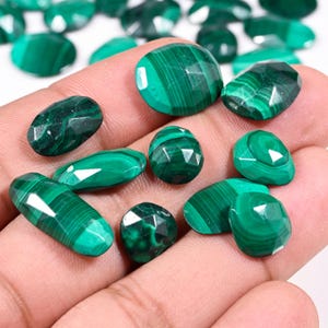 May include: A selection of polished malachite gemstones in diverse shapes, such as oval, round, and faceted. The stones exhibit a deep green hue with characteristic banded patterns. The gemstones are held in a hand.