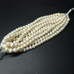 May include: Strands of creamy white pearls, each with a lustrous sheen, are displayed against a dark background. The pearls are strung together, creating a flowing, organic shape. The pearls vary slightly in size, adding to their natural appeal.