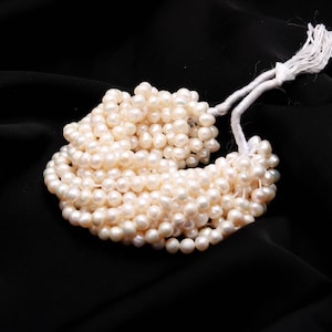 May include: A bundle of creamy white pearls, strung together with a white cord, against a black background. The pearls are small and round, with a slightly irregular shape. The cord is tied at the top with a tassel.