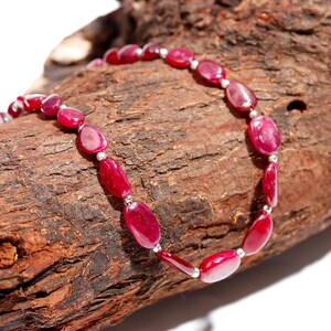 Puede incluir: Un collar con piedras preciosas ovaladas de color rojo intenso, ensartadas con pequeñas cuentas plateadas. Las piedras pulidas tienen un aspecto translúcido. El collar se exhibe sobre un trozo de madera marrón texturizada.