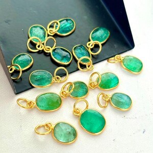 May include: A collection of oval-shaped emerald pendants with gold-colored settings. The pendants are a vibrant green color and have a loop at the top for attaching to a chain or other jewelry component. The pendants are displayed on a dark surface.