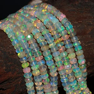 May include: Strands of iridescent opal beads. The beads are a mix of pale blue, green, and pink, with a faceted cut that catches the light. The beads are strung together, creating a shimmering effect. The background is a dark, textured surface.