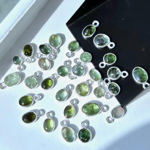 Könnte beinhalten: Eine Sammlung grüner Edelsteinanhänger mit silberfarbenen Schlaufen. Die Edelsteine haben verschiedene Formen, darunter oval und rund, und weisen Grüntöne auf. Die Anhänger sind auf einer weißen Oberfläche mit einem schwarzen, abgewinkelten Hintergrund angeordnet.