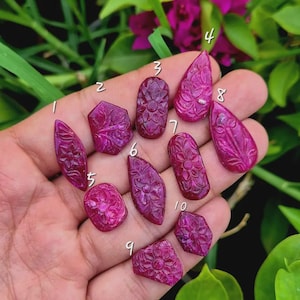 May include: Ten vibrant magenta-colored gemstones, each featuring intricate floral carvings. The stones are numbered one through ten and display various shapes including teardrop, oval, and geometric forms with detailed petal patterns.