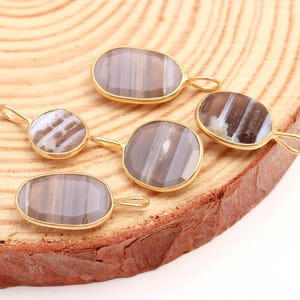 May include: Five gemstone pendants with gold-colored settings. The gemstones are oval and faceted, displaying a mix of gray, white, and brown tones. Each pendant has a small loop for attaching to a chain or other jewelry component. The pendants are arranged on a wooden surface.