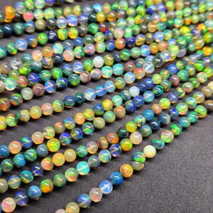 Puede incluir: M&uacute;ltiples hilos de cuentas de &oacute;palo redondas y pulidas que presentan un efecto de juego de colores con destellos en verde, azul, naranja y amarillo. Las cuentas est&aacute;n dispuestas en l&iacute;neas paralelas, mostrando una superficie lisa, reflectante y transl&uacute;cida.