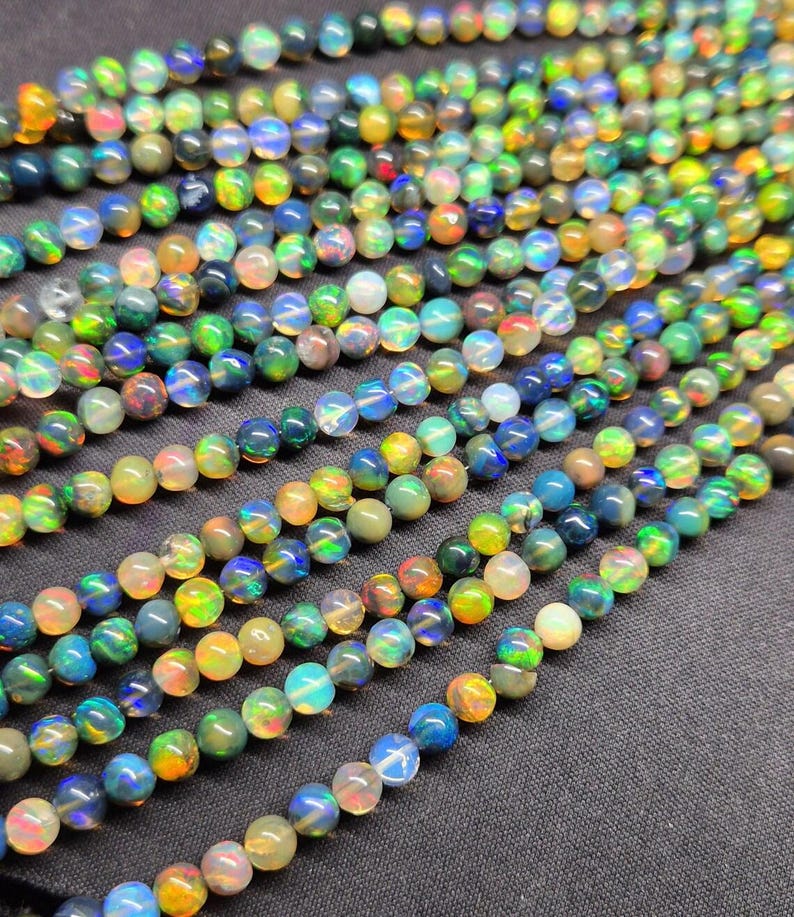Puede incluir: M&uacute;ltiples hilos de cuentas de &oacute;palo redondas y pulidas que presentan un efecto de juego de colores con destellos en verde, azul, naranja y amarillo. Las cuentas est&aacute;n dispuestas en l&iacute;neas paralelas, mostrando una superficie lisa, reflectante y transl&uacute;cida.