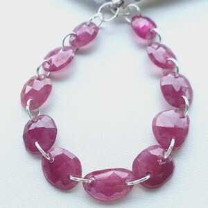 May include: A strand of faceted, oval-shaped, deep pink gemstones, strung together with silver-colored metal links. The gemstones are arranged in a loop, with a white and silver tassel at the top.