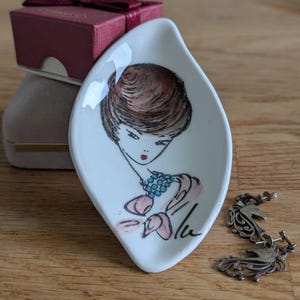 May include: A white, leaf-shaped ceramic dish with a hand-drawn illustration of a woman with brown hair, a blue necklace, and pink accents. A small maroon gift box and silver earrings are also present.