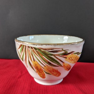 May include: A white ceramic bowl with a gold rim and a floral design featuring yellow and brown leaves. The bowl is sitting on a red surface.