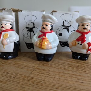 May include: Three ceramic chef figurines in white coats, black pants, and red scarves. Each chef has a white hat and a mustache. One holds a spoon, another a wrapped gift, and the third a checkered napkin. Boxes with chef illustrations are in the background.