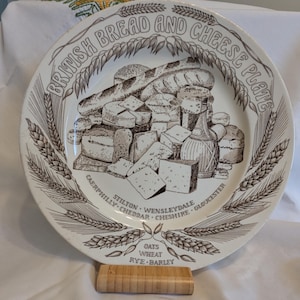 May include: A white ceramic plate with a brown illustration of bread and cheese. The plate is decorated with wheat stalks and the text "BRITISH BREAD AND CHEESE PLATES". Various cheese and bread types are listed around the plate.
