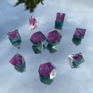 May include: A collection of ten polyhedral dice, featuring a clear, teal, and purple colour scheme. The dice are crafted from resin and have a shimmering, iridescent finish. The numbers are white. These dice are designed for tabletop gaming.