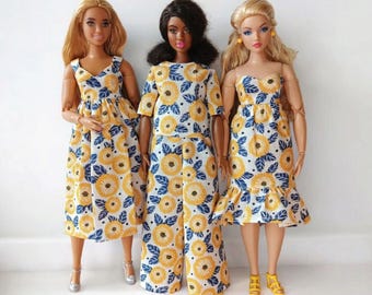 May include: Three dolls wearing matching yellow and blue floral print dresses and jumpsuit. The dolls have different skin tones and hairstyles. One doll wears a v-neck dress, another a jumpsuit, and the third a strapless dress with a ruffled hem.