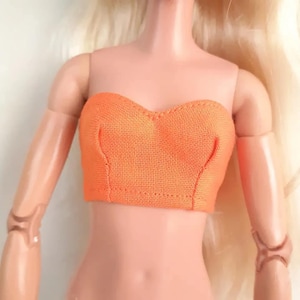 May include: An orange strapless top made of a textured fabric. The top is designed to fit a doll and has a sweetheart neckline. The doll's arms and torso are visible, with blonde hair in the background.
