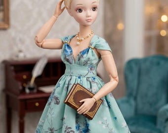 May include: A doll with blonde hair styled in an updo with pearl and crystal accents, wearing a light blue dress with a blue floral pattern. The doll features a pearl necklace and holds a small brown book with gold detailing in its hands.