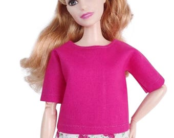 May include: A doll wearing a magenta short-sleeved top and floral print shorts. The doll has blonde hair and is looking forward. The shorts have a white background with pink and green floral patterns.