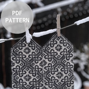 Op de afbeelding: Een paar zwart-witte gebreide vingerloze handschoenen met een geometrisch patroon, hangend aan een waslijn met een houten wasknijper. De handschoenen bevinden zich op een besneeuwde achtergrond. De tekst "PDF PATTERN" is zichtbaar in een witte cirkel.