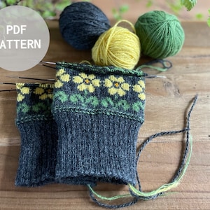 Könnte beinhalten: Ein Paar dunkelgraue Stricksocken mit einem gelben und grünen Blumenmuster. Die Socken liegen auf einer Holzoberfläche mit Wollknäueln in Grün, Gelb und Schwarz.