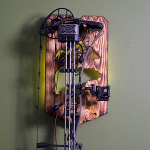 May include: A compound bow mounted on a wooden plaque with a bear silhouette cutout. The plaque has a natural wood grain finish with yellow accents. The bow is black and silver.