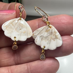 May include: A pair of earrings with a white, fan-shaped design and gold detailing. Each earring has a gold hook and a small, teardrop-shaped charm. The earrings are held up by a person's fingers.