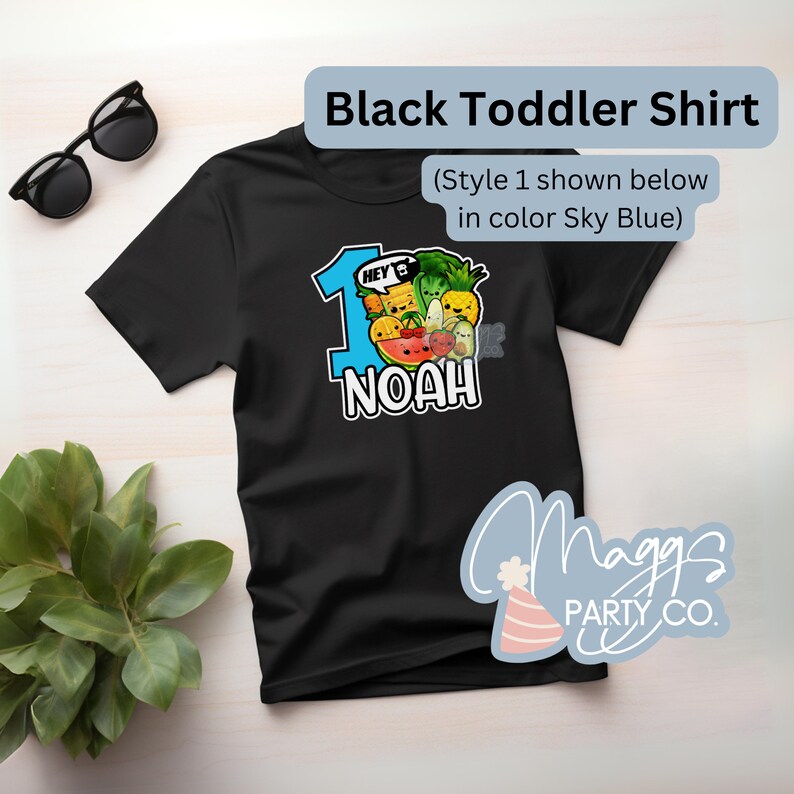 Op de afbeelding: Zwart shirt voor peuters met een blauwe en witte afbeelding van het nummer &eacute;&eacute;n met fruitfiguren en de naam "NOAH". Het shirt wordt getoond op een houten oppervlak met een zonnebril en een potplant.