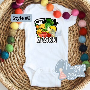 Op de afbeelding: Witte baby romper met een kleurrijk cartoonontwerp van fruit en groenten. Het ontwerp bevat de tekst "Hey 1" en de naam "MASON".