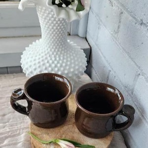 以下が含まれることがあります： ユニークなハンドルデザインの2つの茶色のセラミックマグが木製のコースターに置かれています。背景には花が入った白いホブネイルの花瓶があります。マグカップは空で、光沢のある仕上がりです。1つの花がコースターの上にあります。