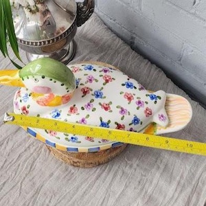 May include: A ceramic duck-shaped serving dish with a white base, adorned with colorful floral patterns. The duck's head is green, yellow, and pink. The dish measures approximately 10 inches in length, perfect for serving appetizers or snacks.
