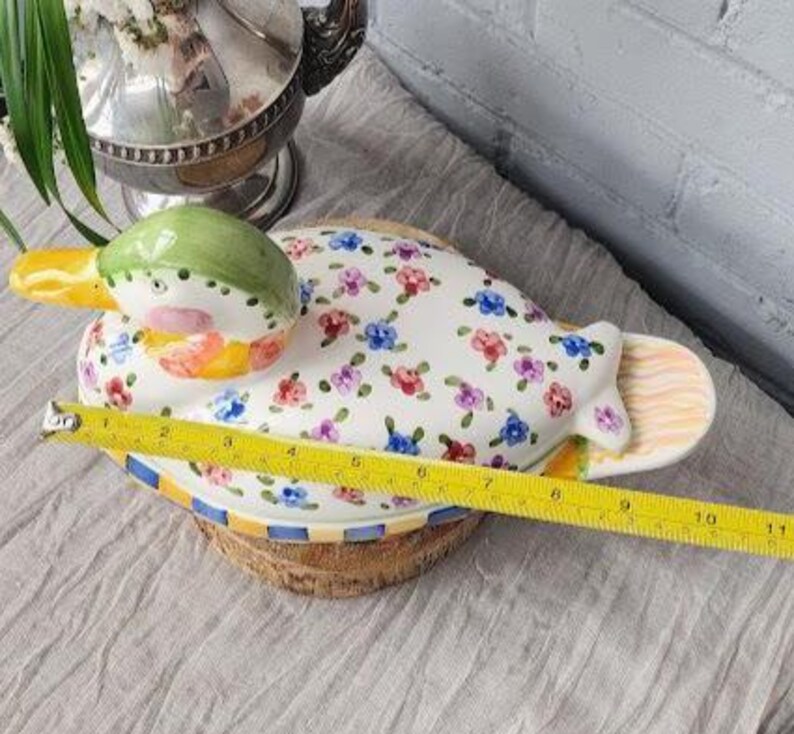 May include: A ceramic duck-shaped serving dish with a white base, adorned with colorful floral patterns. The duck's head is green, yellow, and pink. The dish measures approximately 10 inches in length, perfect for serving appetizers or snacks.