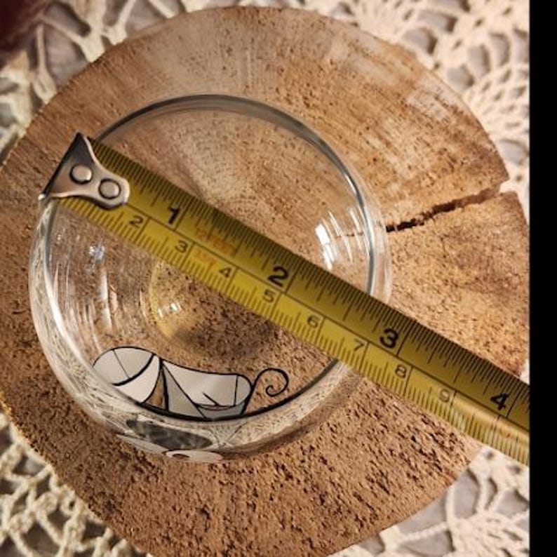 Puede incluir: Un cuenco de vidrio transparente con un dise&ntilde;o de gato en blanco y negro, de aproximadamente 7.6 cm de di&aacute;metro, se mide con una cinta m&eacute;trica amarilla. El cuenco est&aacute; sobre una rodaja de madera redonda y natural.
