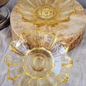 May include: Two matching amber-colored glass candle holders featuring a scalloped, flower-like petal design. Each piece has a central circular depression for a candle, with translucent yellow glass radiating outward in a textured, fluted pattern.
