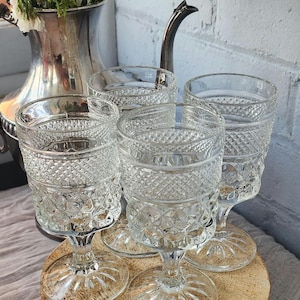 Puede incluir: Cuatro copas de agua transparentes de estilo vintage con cristal con diseño de diamantes. Las copas están sobre una superficie de madera, con una jarra plateada y flores en el fondo. Las copas miden unos 13 cm de alto.