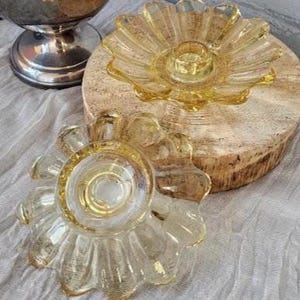 May include: Two yellow glass candle holders with a scalloped, petal-like edge design. One holder is positioned upright on a circular wooden base, while the second holder is placed in front, showing its underside with a central circular indentation.