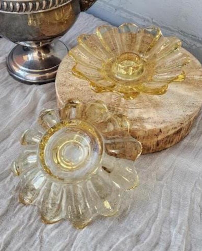 May include: Two yellow glass candle holders with a scalloped, petal-like edge design. One holder is positioned upright on a circular wooden base, while the second holder is placed in front, showing its underside with a central circular indentation.