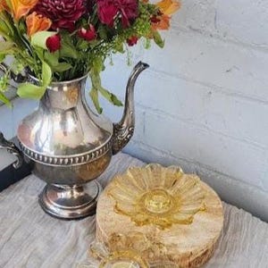 May include: A silver-toned metal teapot filled with a bouquet of orange, red, and purple flowers, accompanied by two amber-colored glass candle holders with a scalloped, petal-like design, one resting on a circular wooden base.
