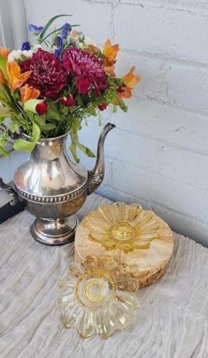 May include: A silver-toned metal teapot filled with a bouquet of orange, red, and purple flowers, accompanied by two amber-colored glass candle holders with a scalloped, petal-like design, one resting on a circular wooden base.