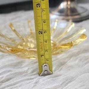 May include: A clear and amber-colored glass bowl with a scalloped, petal-like edge. A yellow measuring tape is positioned vertically against the bowl, showing a height of approximately 2.5 inches from the base to the rim of the glass dish.