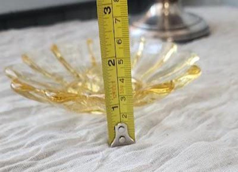 May include: A clear and amber-colored glass bowl with a scalloped, petal-like edge. A yellow measuring tape is positioned vertically against the bowl, showing a height of approximately 2.5 inches from the base to the rim of the glass dish.