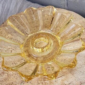 May include: A translucent yellow glass candle holder featuring a scalloped, flower-like edge and a central circular opening designed to hold a taper candle. The glass has a smooth, reflective surface with a distinct radial pattern radiating outward.